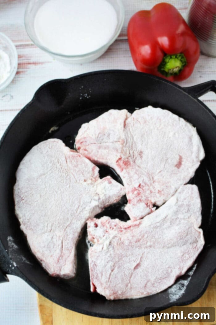 Tropical Pork Chops 4 Three flour-coated raw pork chops in a black skillet, with a red bell pepper and a bowl nearby—perfect ingredients for trying new pork chop recipes.