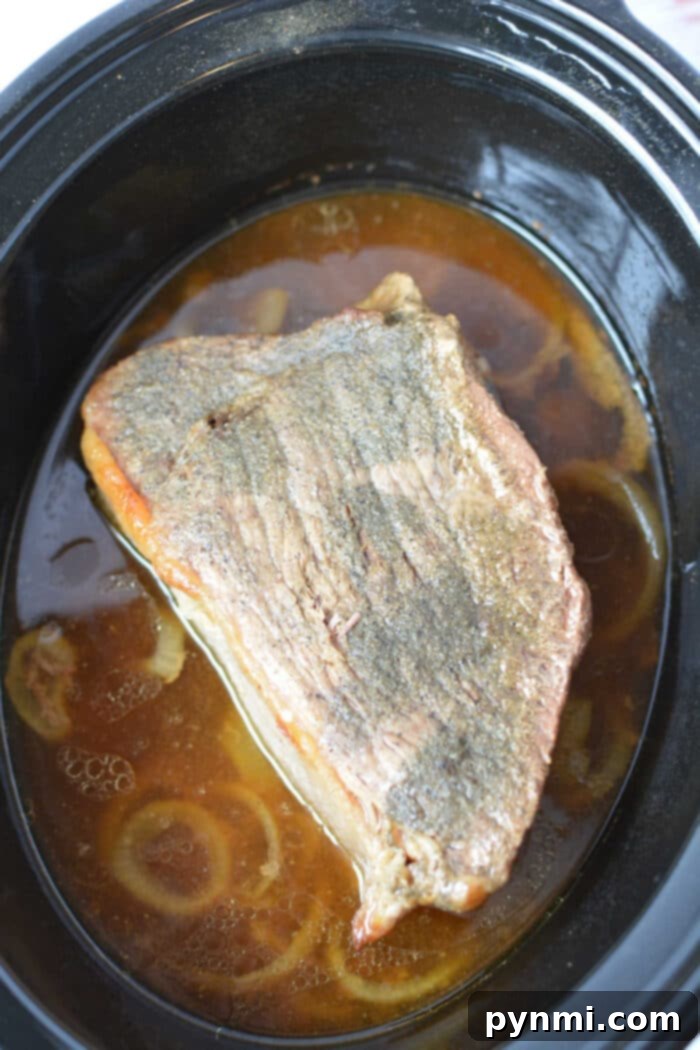 A piece of cooked Beef Bottom Round Roast sits in broth with onions inside a black slow cooker.