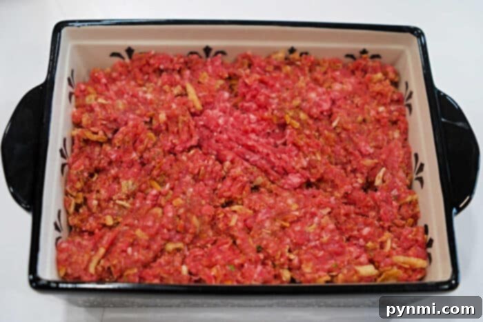 Ultimate BBQ Glazed Meatloaf 5 Raw ground beef, seasoned and mixed with onions, is carefully spread evenly within a rectangular baking dish, preparing for a classic meatloaf recipe or a savory BBQ meatloaf transformation, perfectly prepped for cooking.