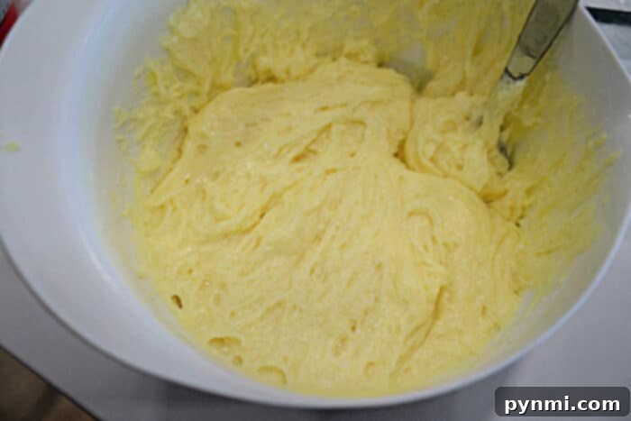 A white bowl filled with creamy, pale yellow lemon angel food cake batter, with a metal spoon partially dipped in, showcasing the smooth, airy consistency.