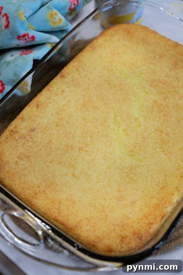 A freshly baked, golden-brown lemon angel food cake in a clear glass baking dish, resting on a cooling rack beside a vibrant yellow and white kitchen towel, inviting a delicious tasting experience.
