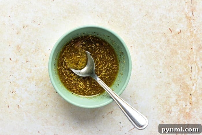 A small blue bowl with herb marinade and a spoon—a perfect accent for oven roasted broccolini—rests on a light textured surface.