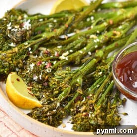 Oven roasted broccolini with lemon wedges and dipping sauce on a plate, sprinkled with sesame seeds and spices.
