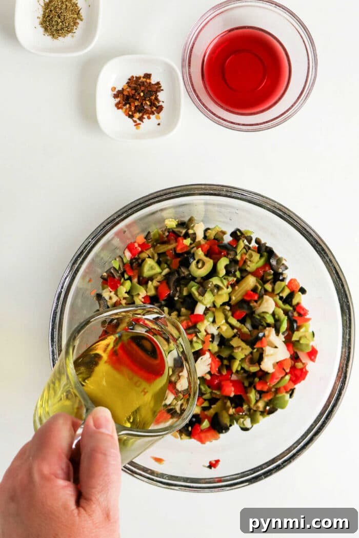 A hand pours olive oil into a bowl of chopped vegetables for a classic Olive Salad, with spices and red vinegar nearby—perfect for an authentic Muffuletta recipe.