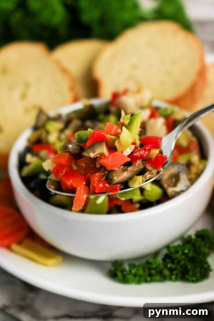 A spoonful of mixed chopped vegetables above a bowl, hinting at a classic Olive Salad recipe, with bread slices in the background.