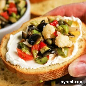 A hand holds a slice of bread topped with cream cheese and a vibrant Olive Salad, featuring chopped olives, cauliflower, and peppers—a perfect bite inspired by the classic Muffuletta recipe.