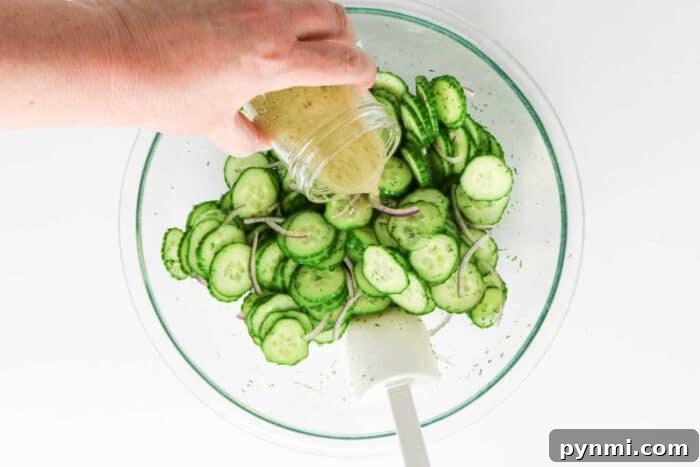 Crisp Cucumber Dill Salad with Creamy Yogurt Dressing 5 A hand pours dressing over sliced cucumber and onion in a glass bowl with a white spatula, creating a fresh salad perfect for adding dill.