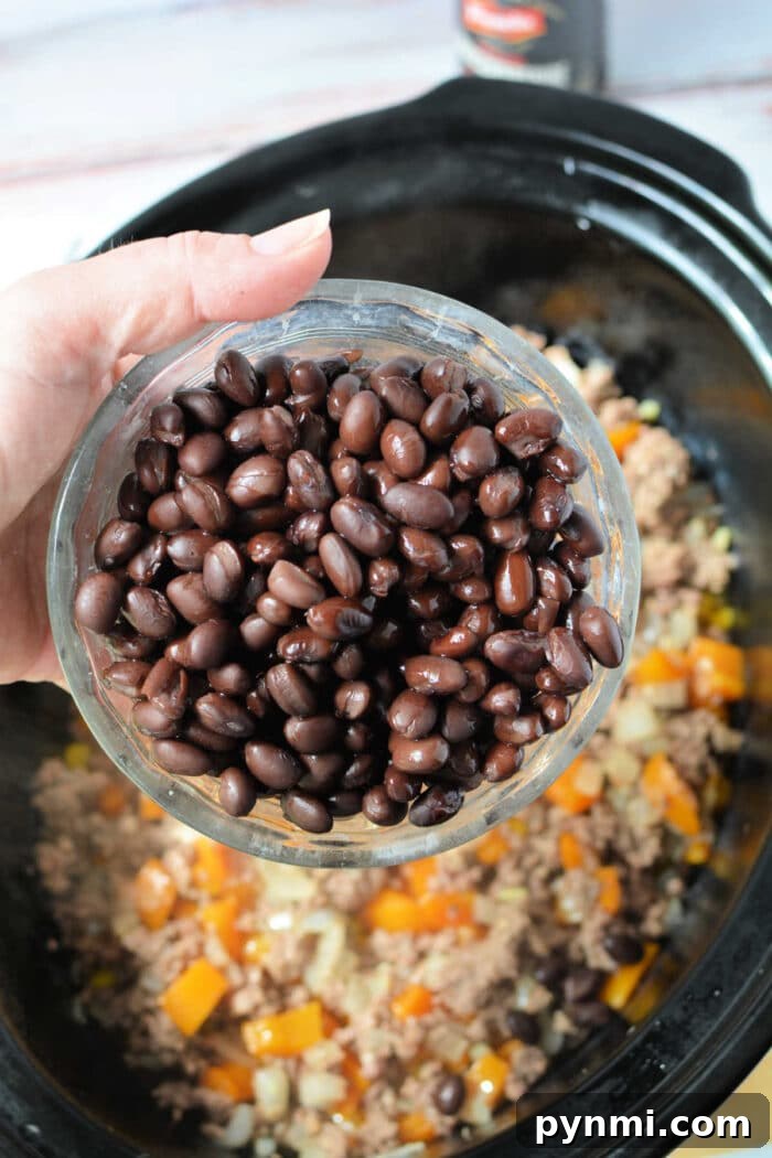 Slow Cooker Cowboy Casserole 4 A hand holds a bowl of black beans over a Crockpot filled with ground meat and chopped vegetables, preparing a hearty Cowboy Casserole.