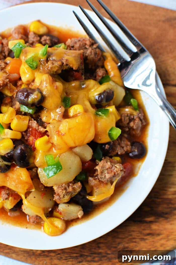 Slow Cooker Cowboy Casserole 6 A plate of cheesy Cowboy Casserole with beef, black beans, corn, and potatoes sits next to a fork—perfect for fans of hearty Crockpot recipes.