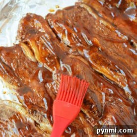 A red brush spreads barbecue sauce over beef riblets on a foil-lined baking sheet, making this the perfect beef riblets recipe for your next cookout.