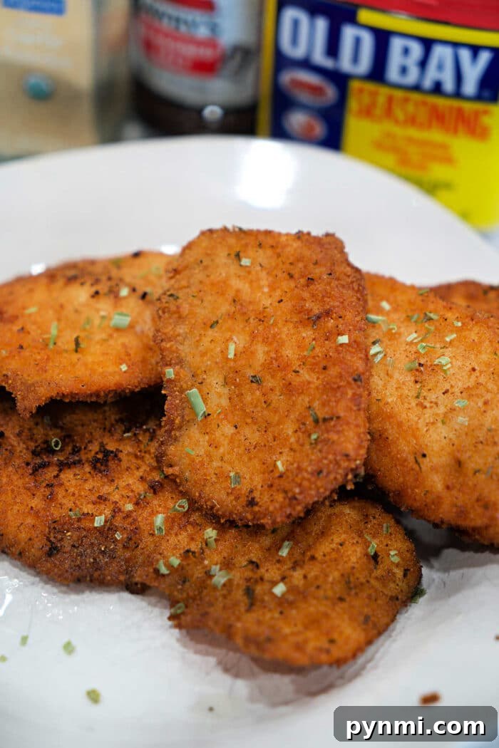 Golden, crispy breaded chicken cutlets on a plate, garnished with herbs, with Old Bay seasoning and a side of fried tilapia for the perfect tilapia recipe in the background.
