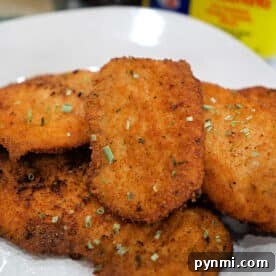 Golden, crispy breaded chicken cutlets on a plate, garnished with herbs, with Old Bay seasoning and a side of fried tilapia for the perfect tilapia recipe in the background.