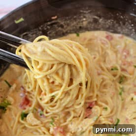 Tongs lifting creamy chicken spaghetti with tomatoes and herbs from a black bowl, fresh from the crockpot.