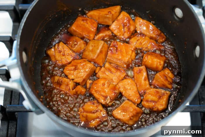 Sticky Teriyaki Salmon Bites Recipe with Sesame Glaze 6 Chunks of Teriyaki Salmon Bites cooking in a dark, glossy sauce in a saucepan on a stovetop.