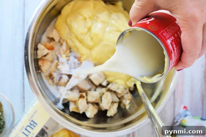 Cheesy Ritz Chicken Casserole with Crispy Cracker Topping 4 A hand pours canned milk into a bowl with diced chicken and creamy yellow sauce, preparing a delicious mixture for Ritz Chicken Casserole.