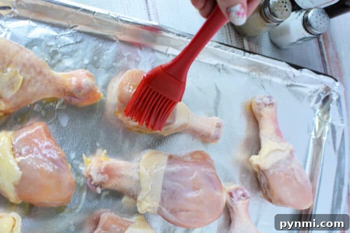 How to Bake Frozen Chicken Legs for Crispy, Juicy Results 4 A hand brushes raw frozen chicken legs with oil on a foil-lined baking sheet, salt and pepper nearby, preparing them perfectly for baked frozen chicken.
