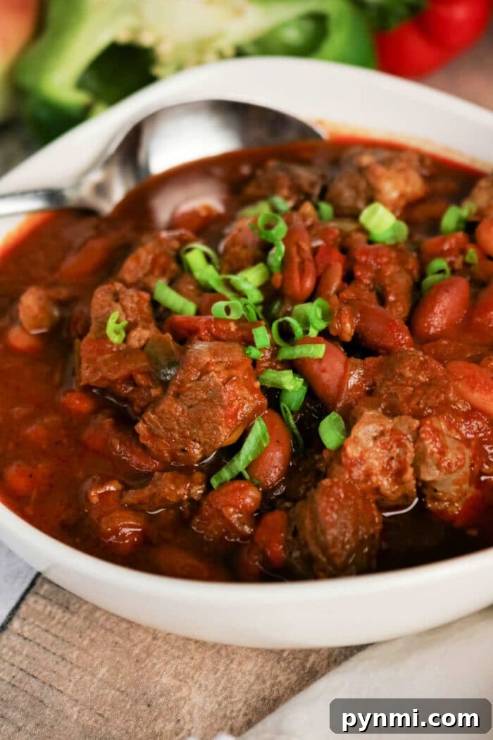 Bowl of hearty Chuck Roast Chili with beef, beans, and chopped green onions, with a spoon resting in the bowl.