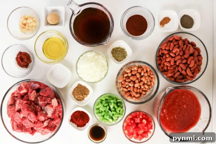 Bowls of raw beef, beans, diced vegetables, spices, and liquids arranged on a white surface, viewed from above—ingredients perfectly suited for a hearty Chuck Roast Chili.