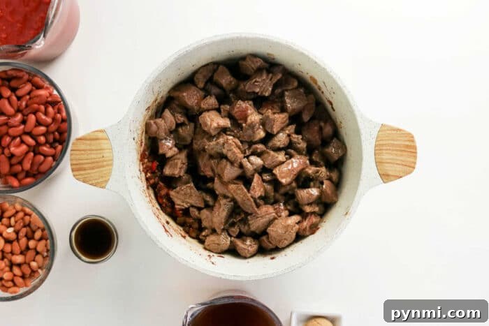 Overhead view of cubed beef in a white pot, perfect for making Chuck Roast Chili, surrounded by beans, sauce, and seasonings on a white surface.