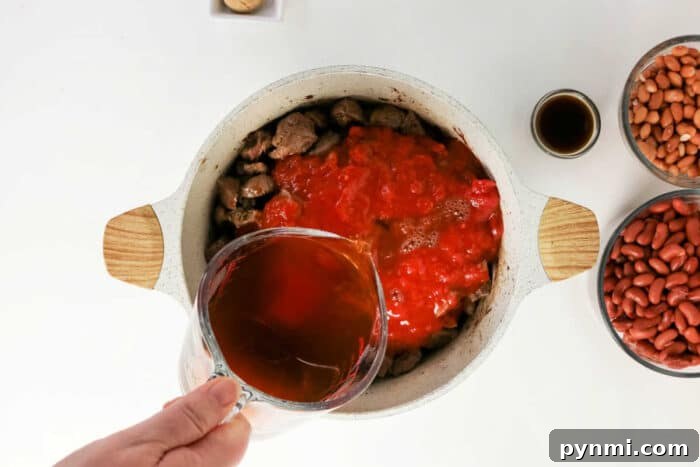 A hand pours broth into a pot with beef and tomatoes, surrounded by beans and sauce on a white table, creating the perfect base for a hearty beef chili recipe.