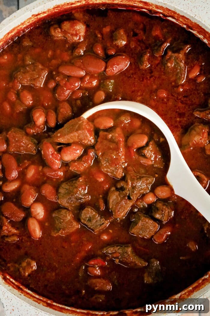 A white ladle scoops rich Chuck Roast Chili, brimming with beef and beans, from a pot filled with thick, red, hearty stew.